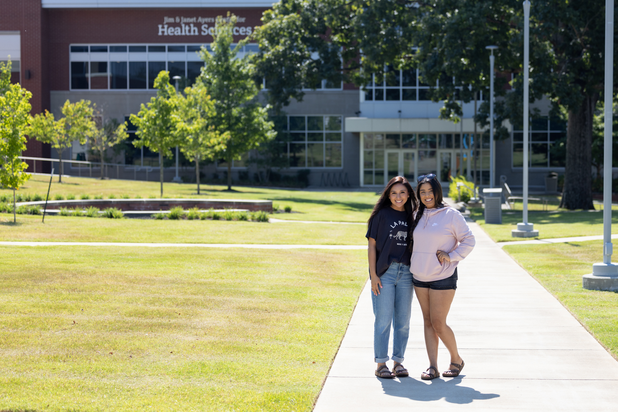 Two female students stand on a sideway on the JSCC Jackson Campus, posed together while smiling at the camera.