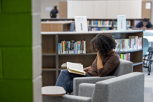 A student reads a book while sitting next to a bookshelf in the newly renovated JSCC Learning Commons.