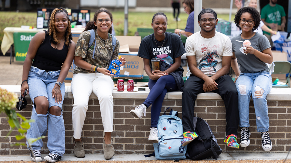 A group of students smile at the camera while sitting in the JSCC quad.