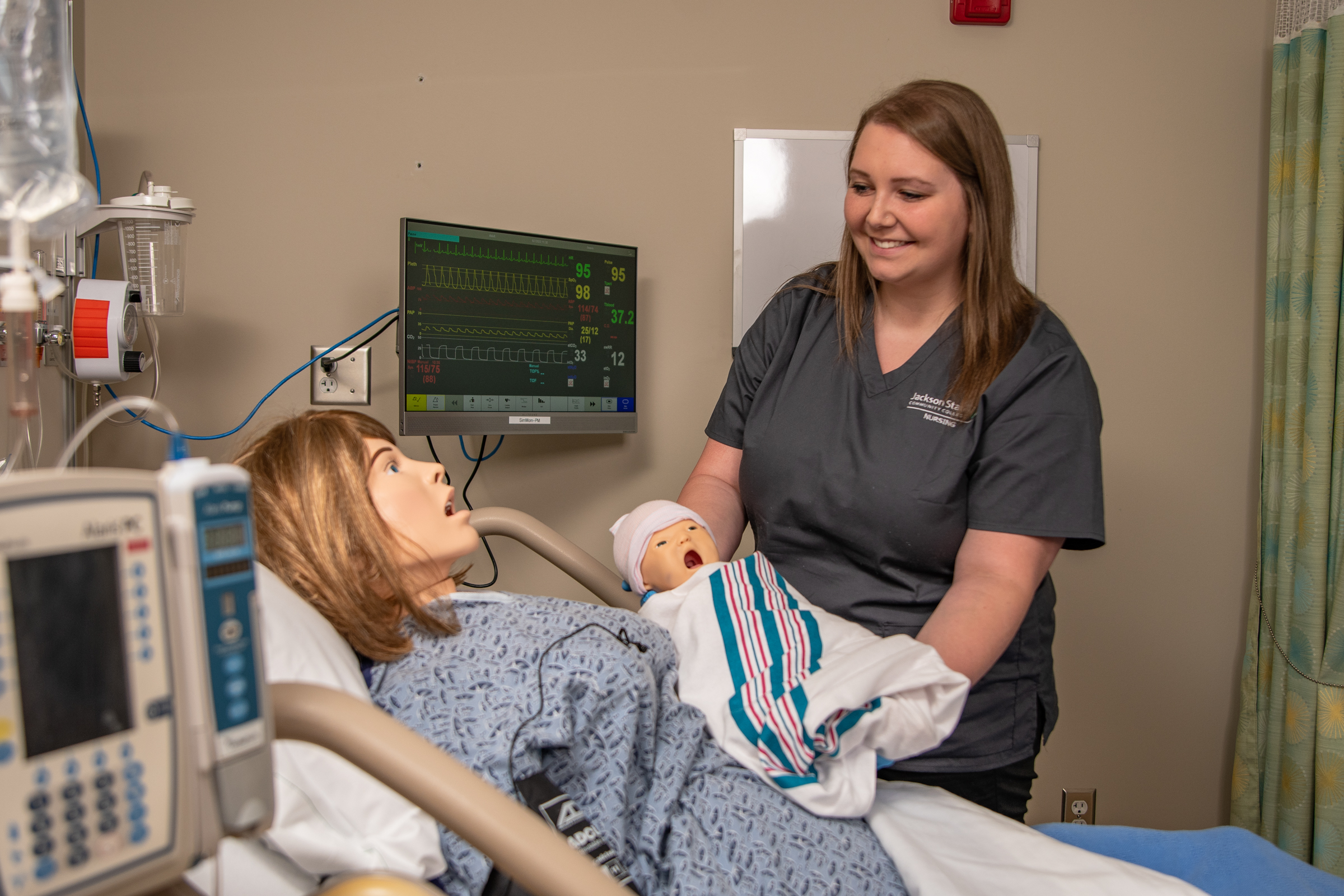 Nursing student handing a simulation baby to it's mother in a hospital simulation room.