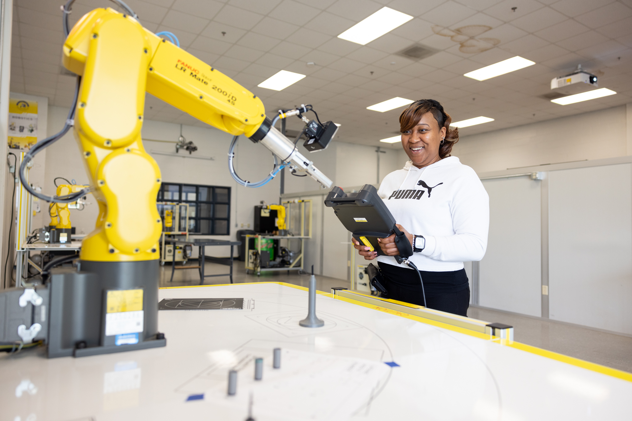 Woman smiling while holding an industrial controller for a FANUC Robot as the robot completes tasks that the woman inputs.