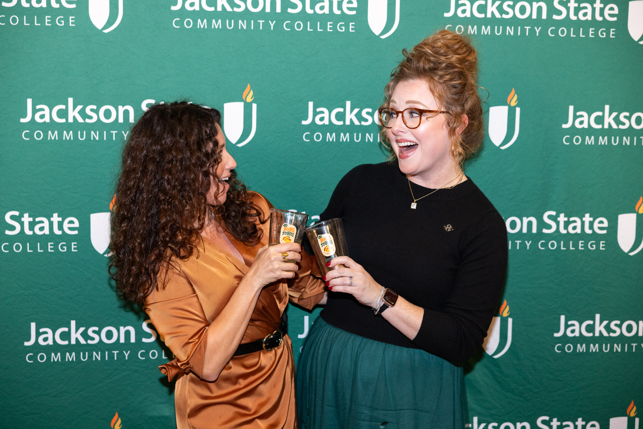 Two women pose for a photo as they toast with pint glasses at a social event called Tacos, Tecates & Scholarships.