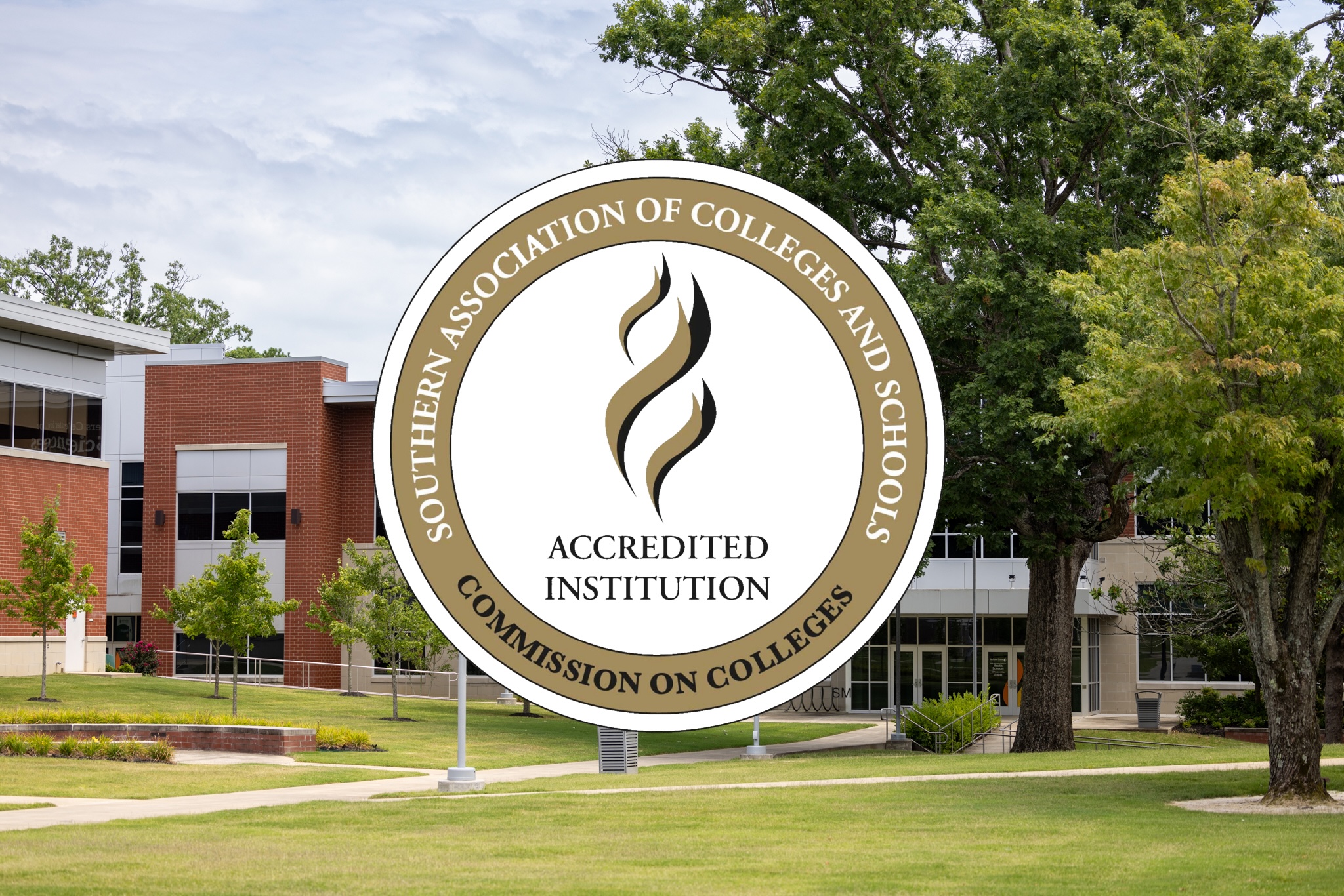 A large circular sign in the foreground displays the logo and text of the Southern Association of Colleges and Schools Commission on Colleges. The text around the outer ring reads “Southern Association of Colleges and Schools Commission on Colleges.” Inside the circle, there is a stylized flame logo above the words “Accredited Institution.” The sign is placed on a photo of Jackson State’s campus.