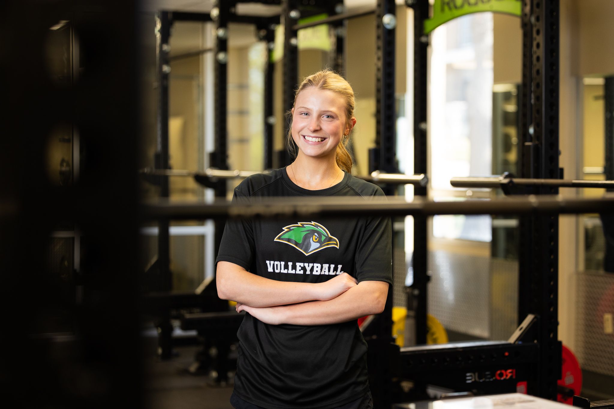 Photo of Emily Papple in the JSCC Fitness Center with arms crossed wearing a JSCC Green Jays Volleyball Workout shirt.
