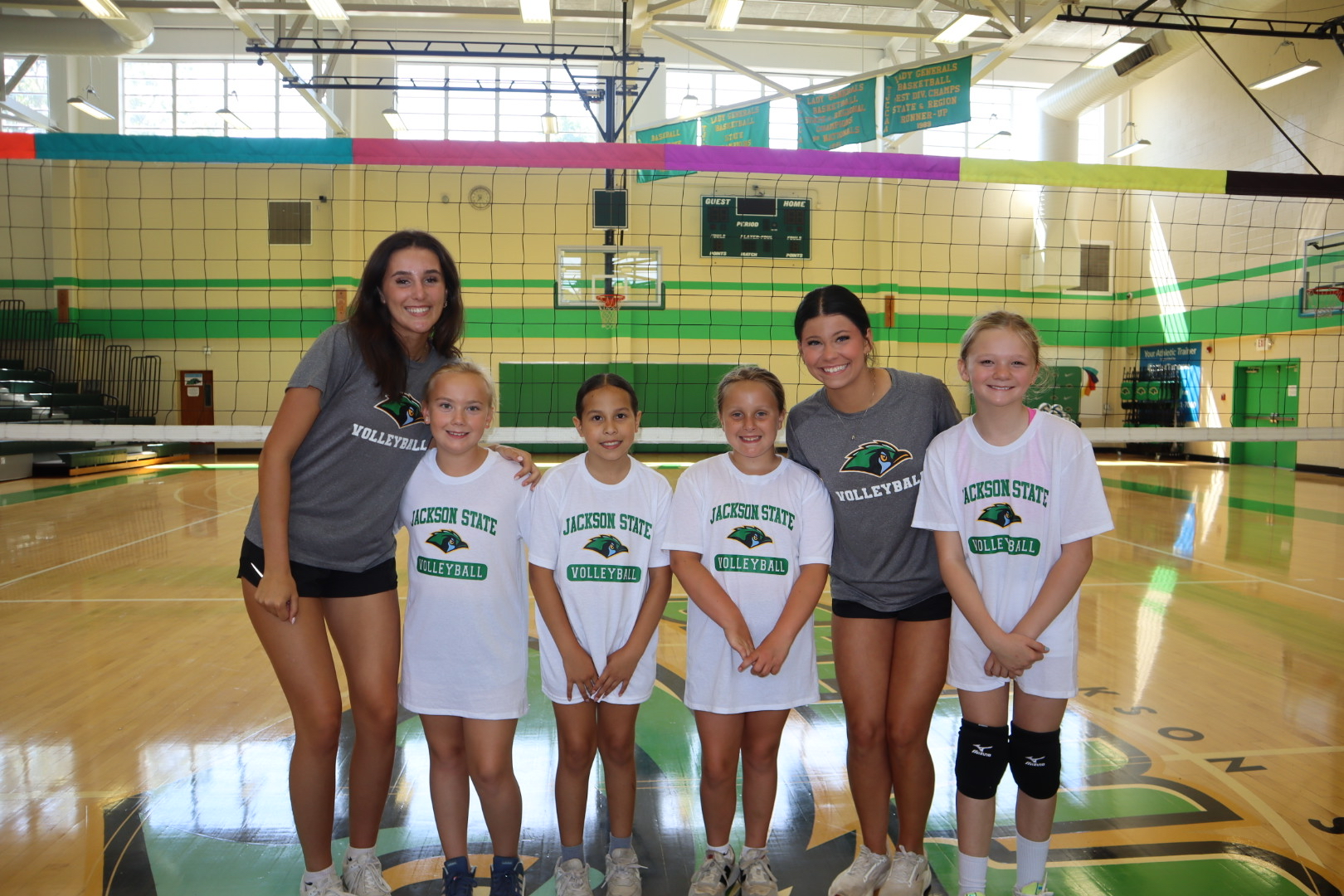 Promo image for Summer Volleyball Camps Series in the JSCC Gymnasium showing two JSCC Volleyball players with four young camp attendees posing together for a photo.